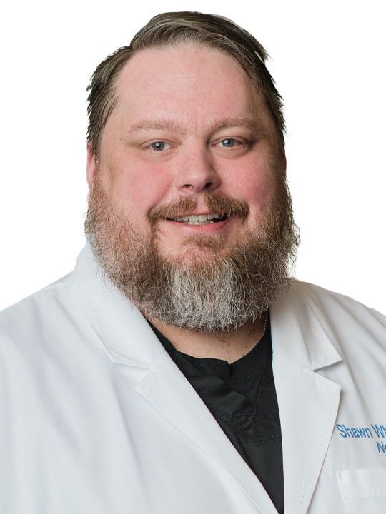 A man with a beard is wearing a white lab coat and smiling.