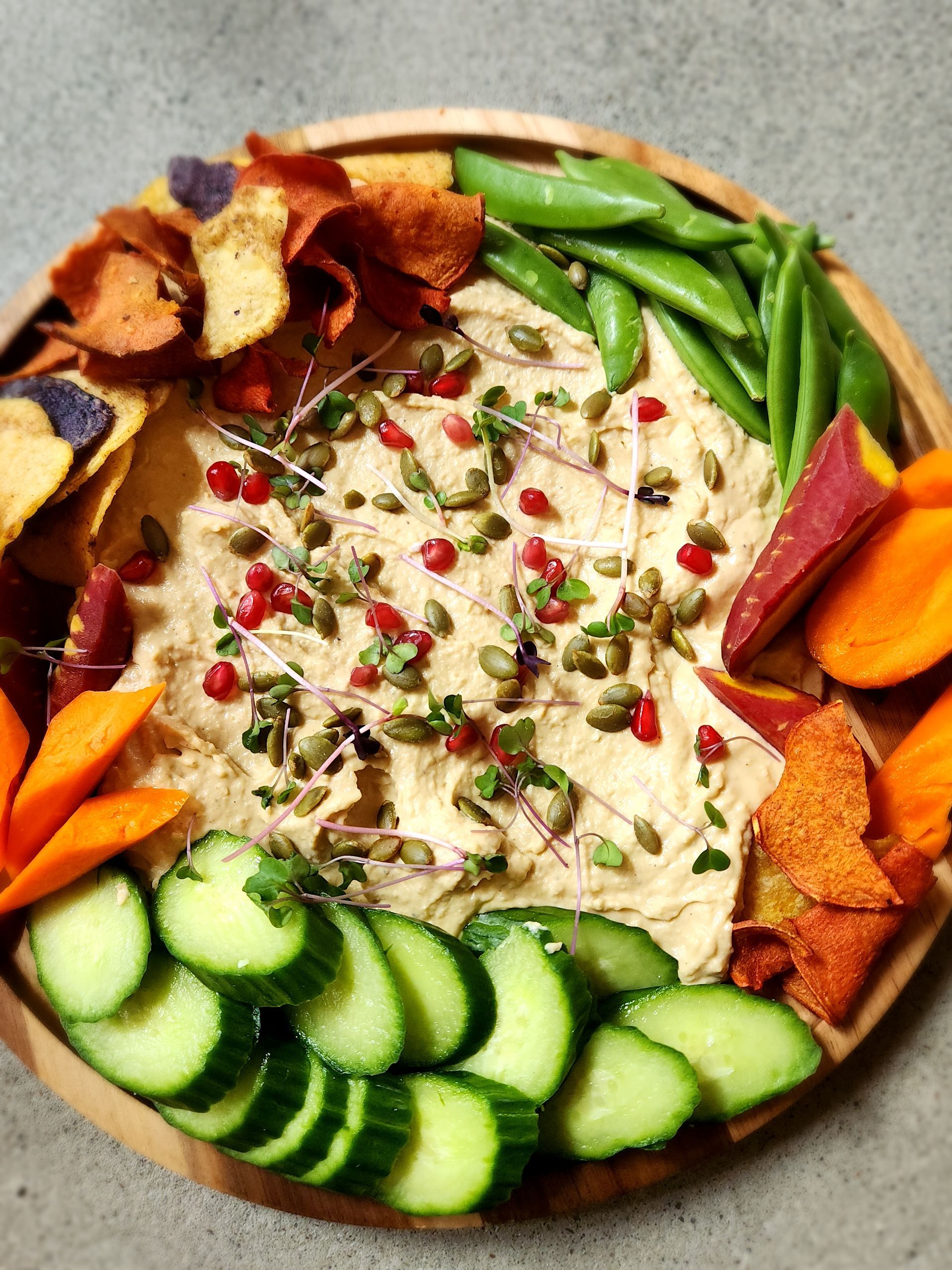 A wooden plate topped with hummus , vegetables and chips.