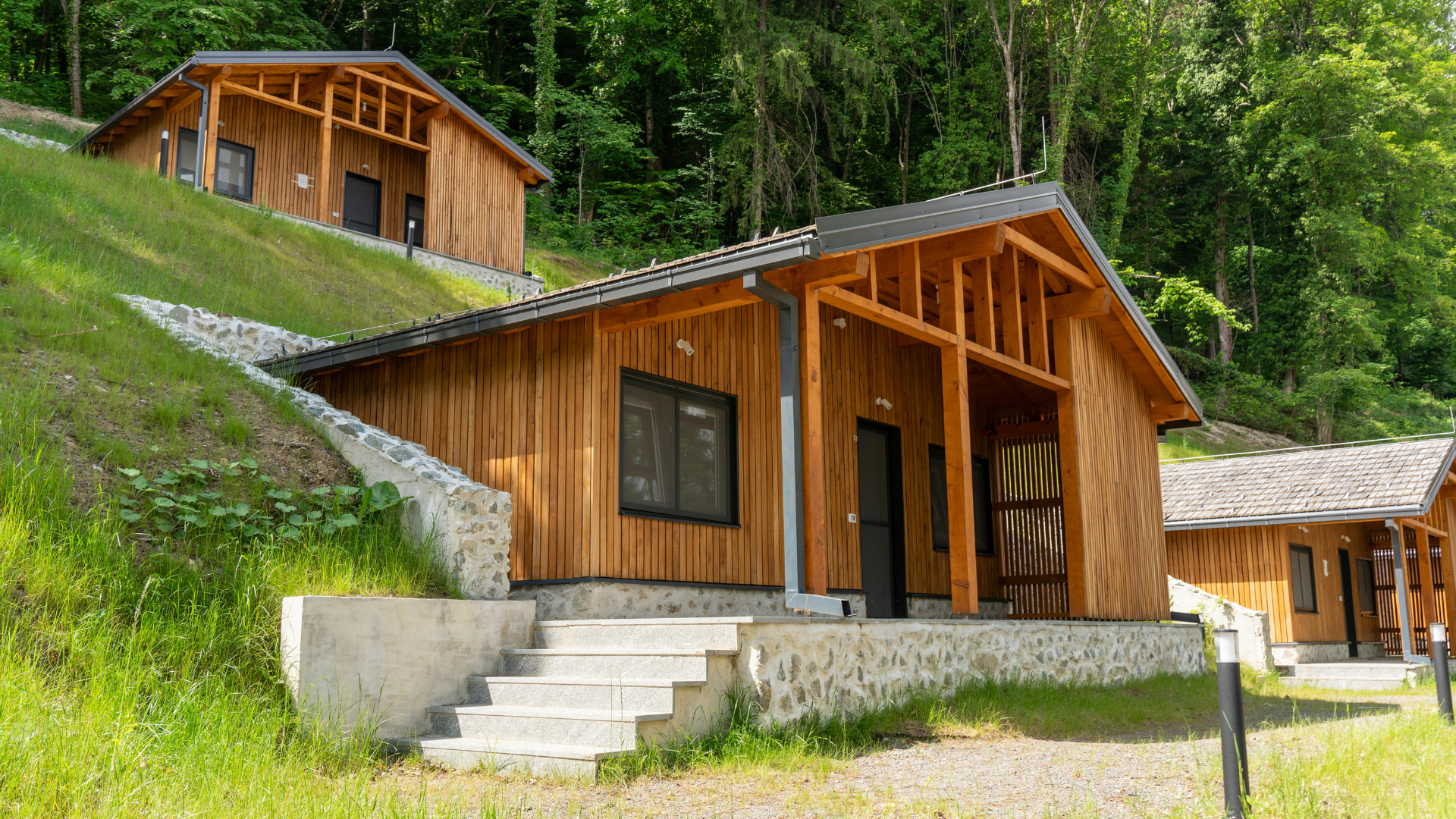 Wooden cabins on a hillside, surrounded by greenery. Each cabin has a dark roof and a porch.