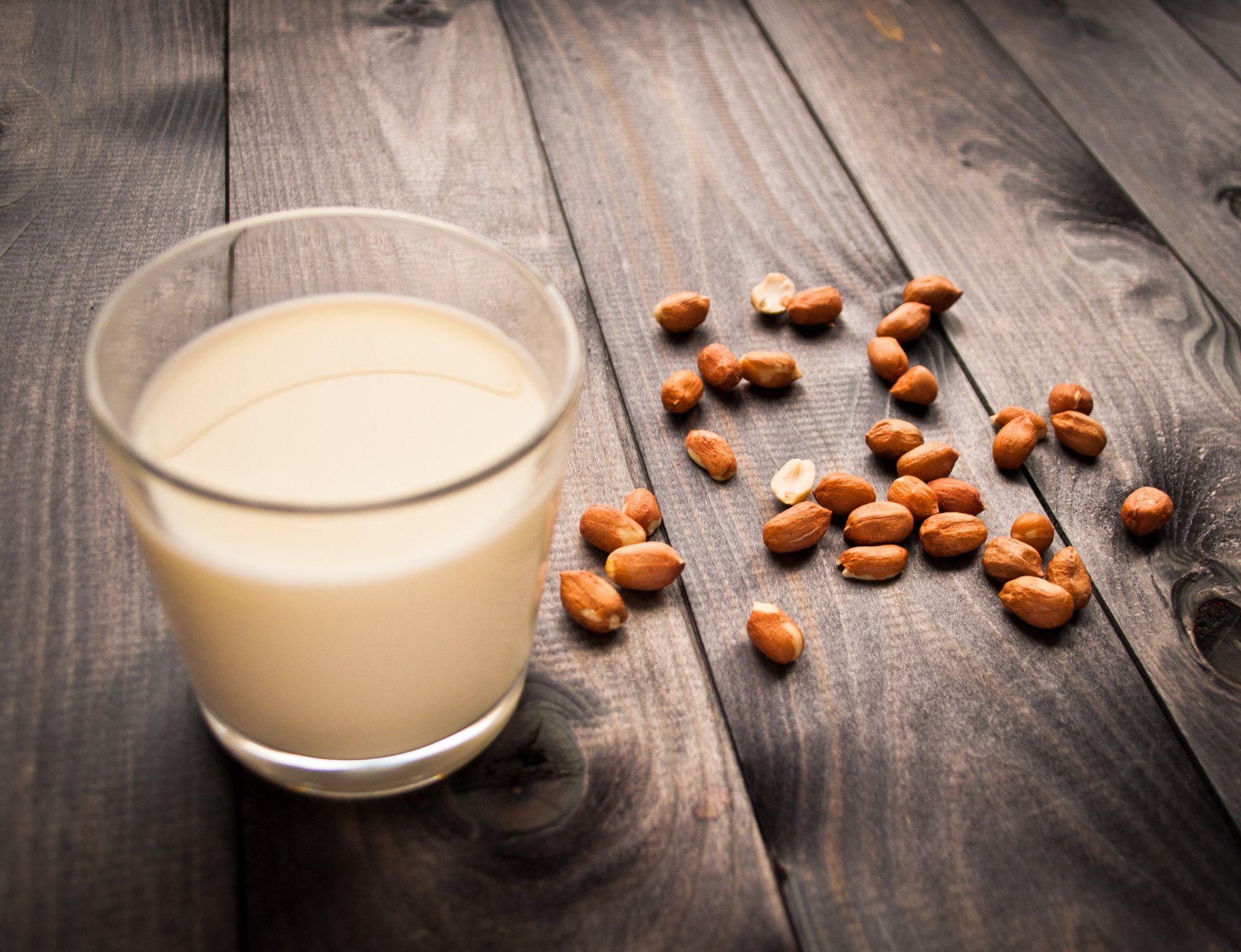 A glass of peanut milk is on a wooden table next to peanuts.