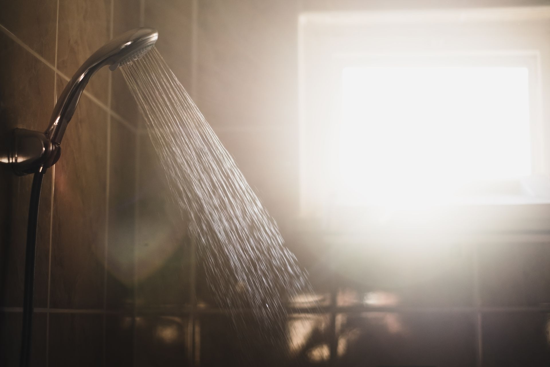 A shower head spraying water against a tiled wall in a dimly lit, sunlit bathroom.