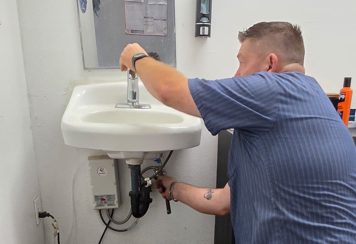 A person in a blue shirt repairs a wall-mounted bathroom sink, using a tool on the plumbing underneath.
