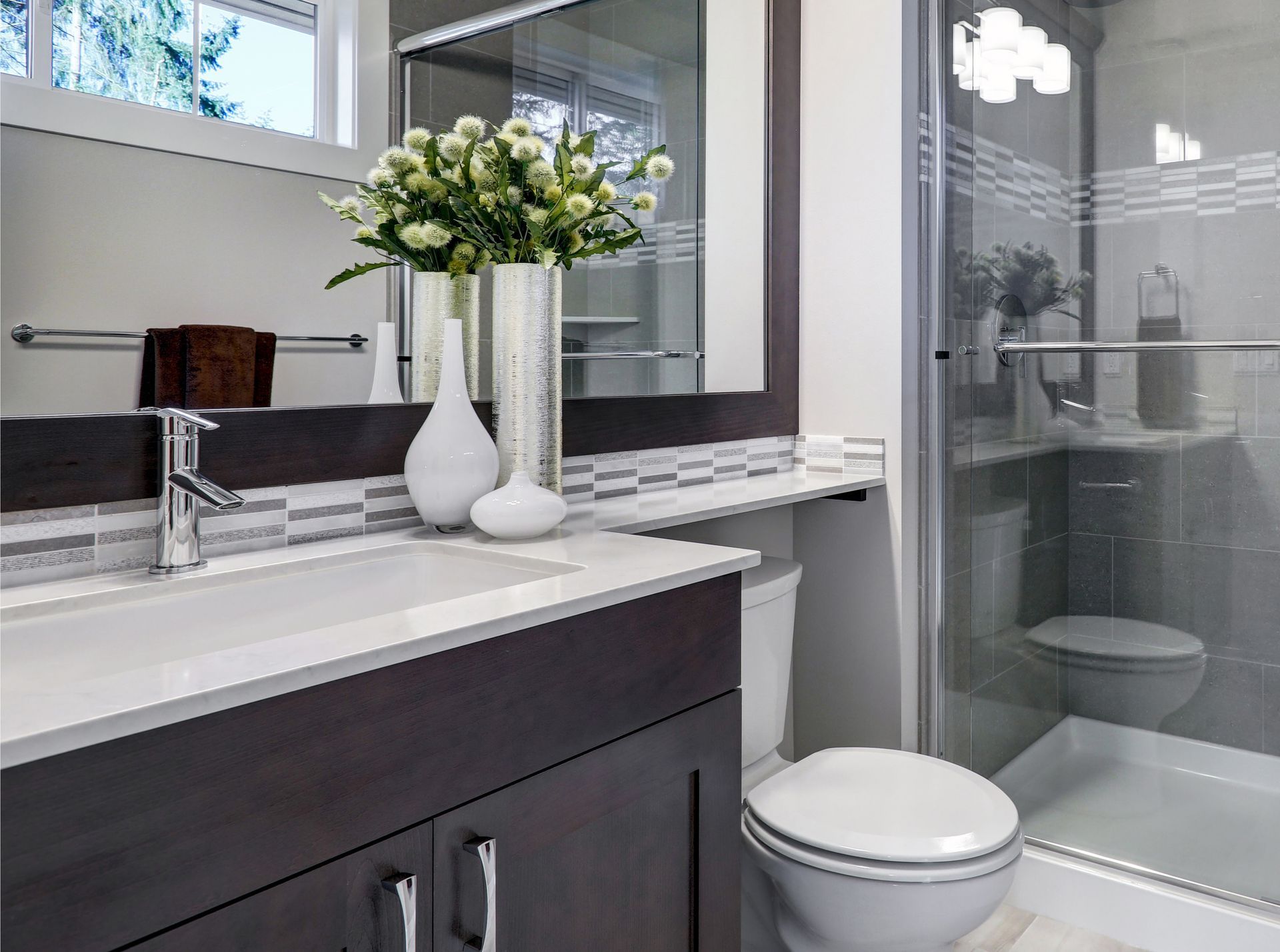 A modern bathroom with a dark wood vanity, white countertop, sink, large mirror, and a glass-enclosed shower.