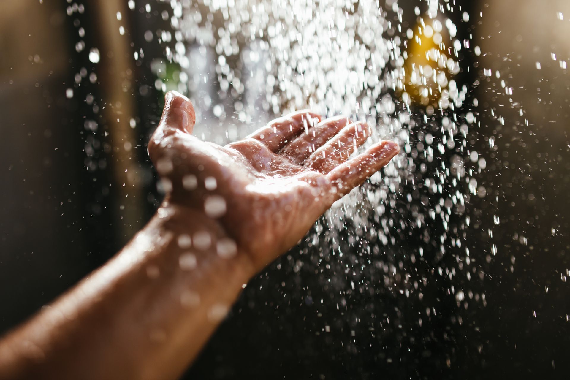 A hand reaches upward into a spray of water droplets, illuminated against a dark background.