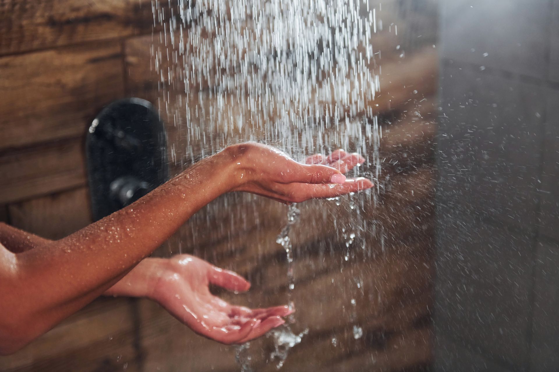 A person’s hands cupped under a stream of water from a shower head against a tiled wall.