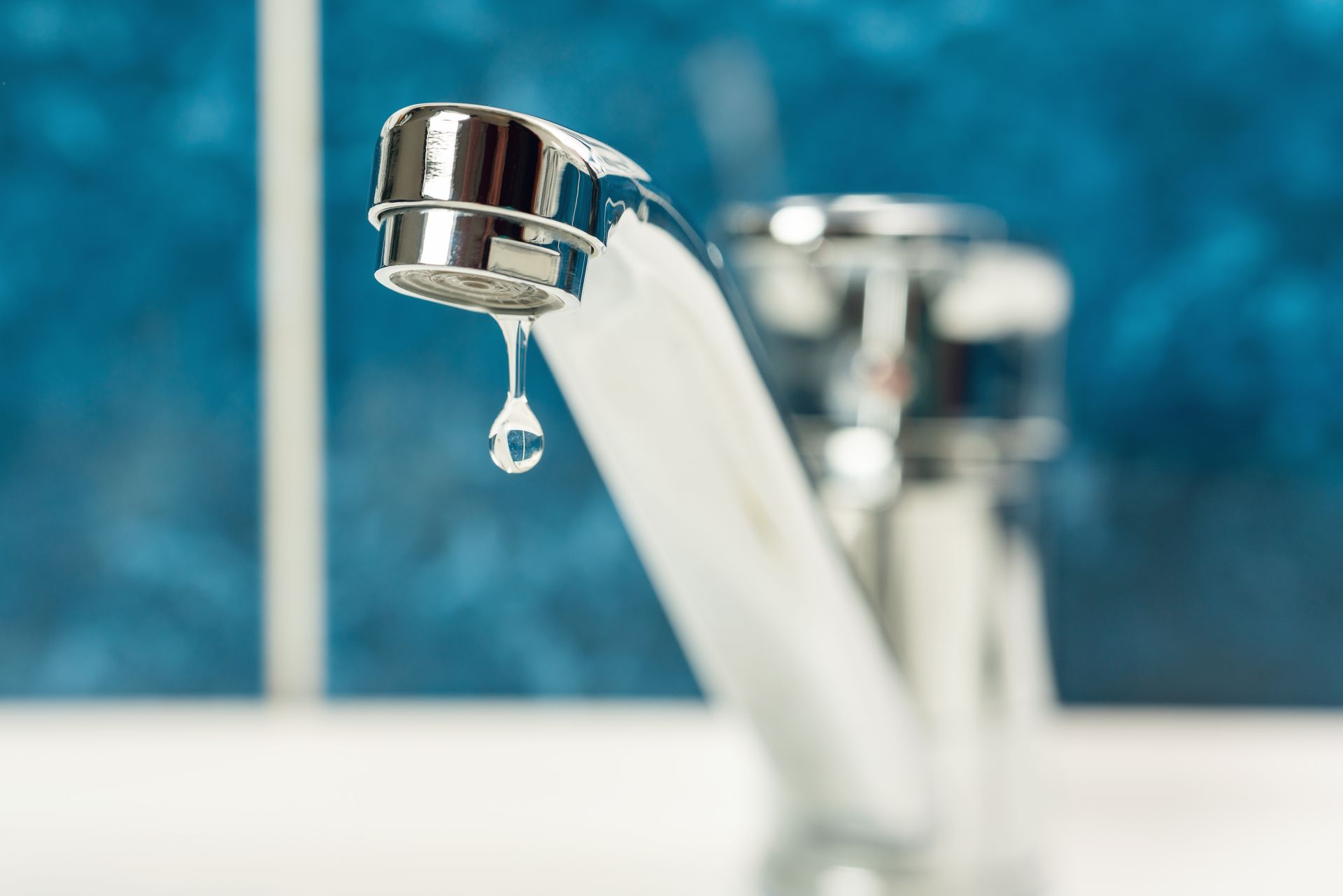 A single drop of water hangs from a chrome faucet against a blurred blue background.