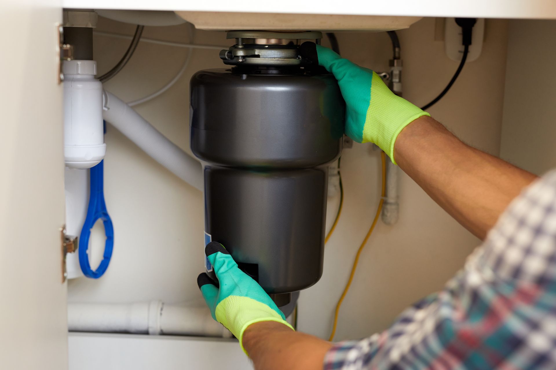 A person wearing lime green work gloves installs a dark gray garbage disposal unit under a kitchen sink.