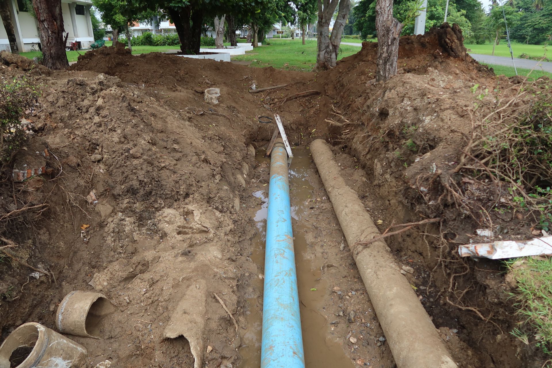 A trench in the ground revealing a blue PVC pipe running parallel to an older concrete pipe.
