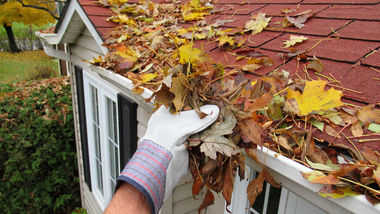 Gloved hand clearing wet leaves from a red shingled roof near a dormer window
