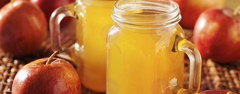 Three mason jars filled with apple juice are sitting on a table next to apples.
