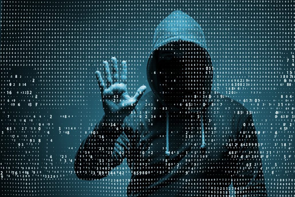 A man in a hoodie is standing in front of a computer screen.