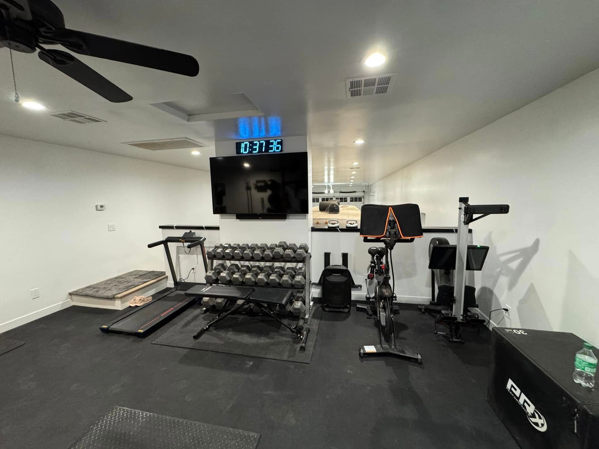 Home gym with various exercise equipment including a treadmill, weight bench, and stationary bike.  Black floor, white walls, and a large TV are visible.
