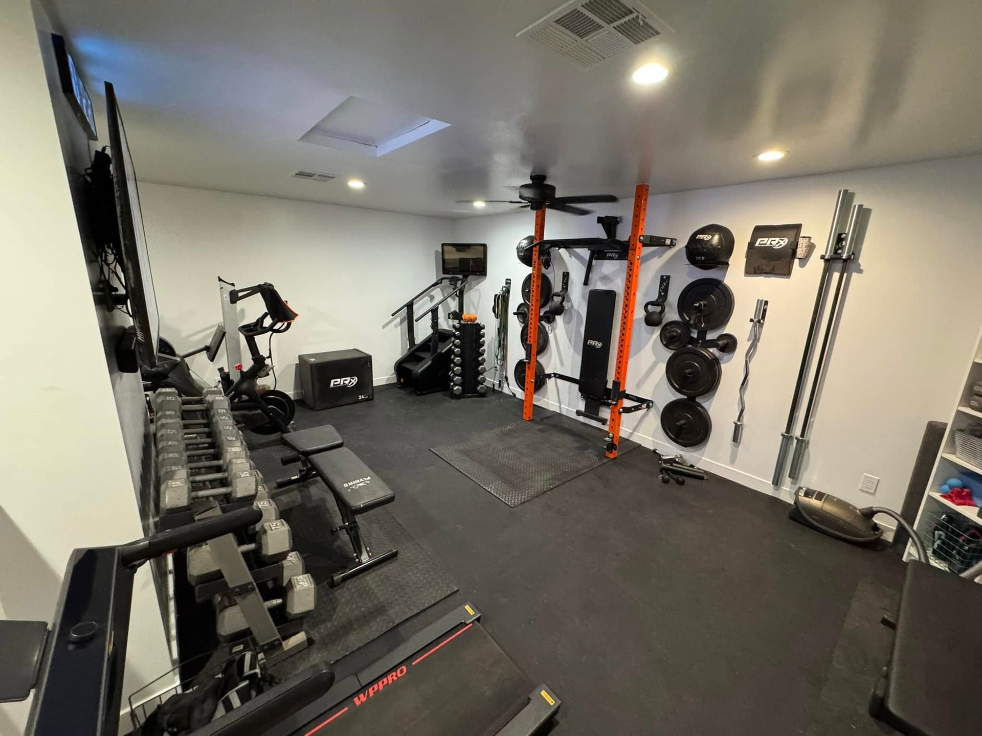 A well-equipped home gym featuring dumbbells, weight plates, barbells, and cardio machines on a rubber floor. The walls are white and equipment is orange and black.