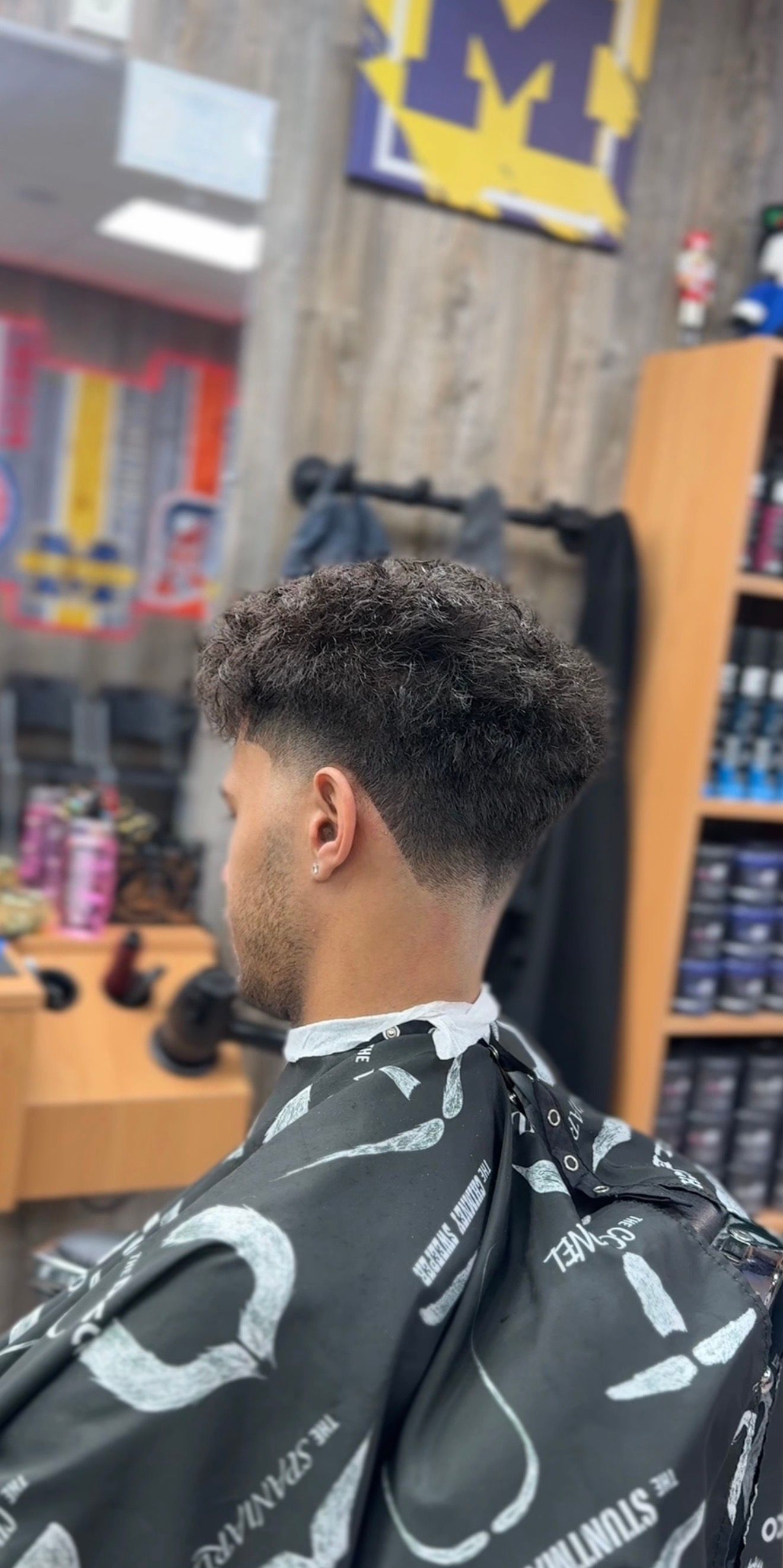 A person getting a haircut in a barbershop. He has curly hair with a fade. The cape is black and white.
