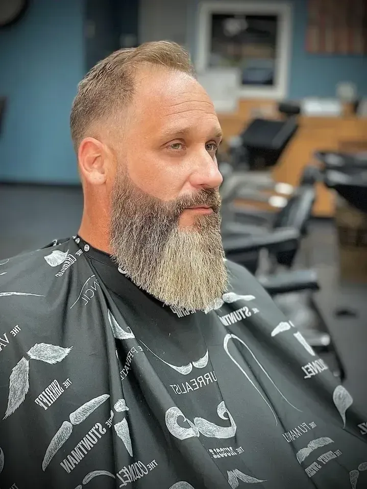 Man with a full beard, fade haircut, wearing barber cape, sitting in a barbershop.