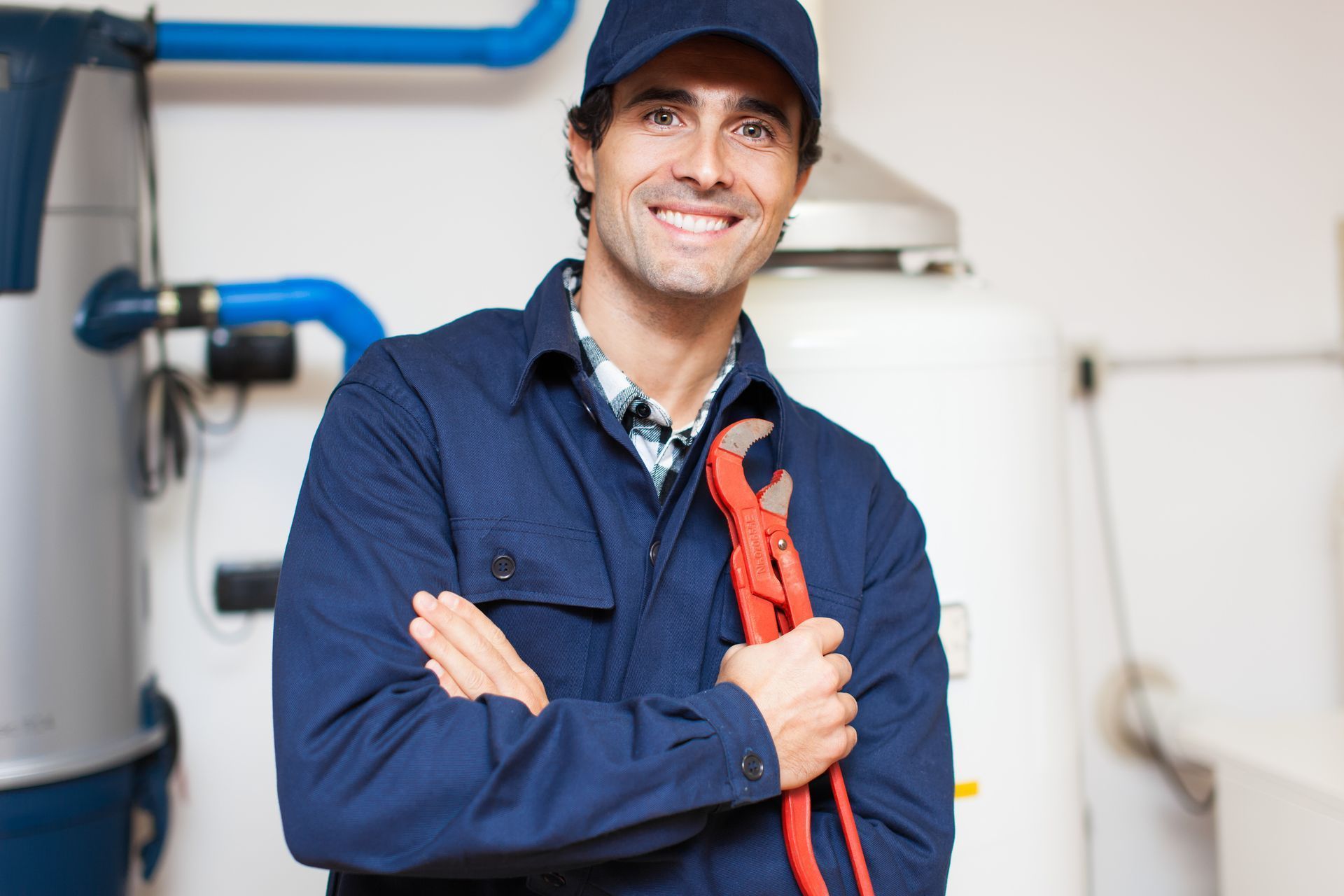 Technician in blue workwear smiling beside plumbing equipment and blue pipes, holding a red wrench