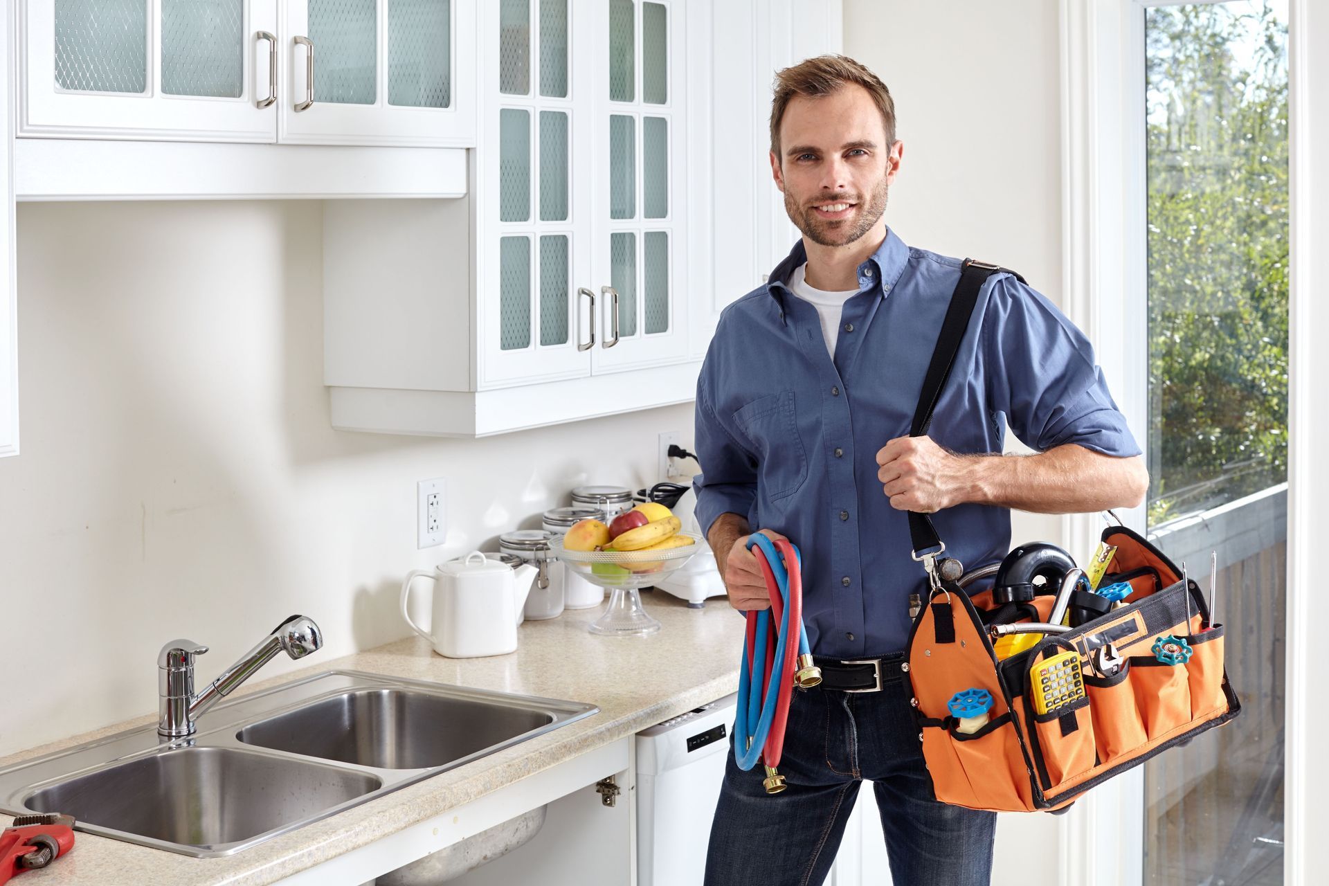 Plumber holding tools beside a kitchen sink and cabinets in a bright home kitchen
