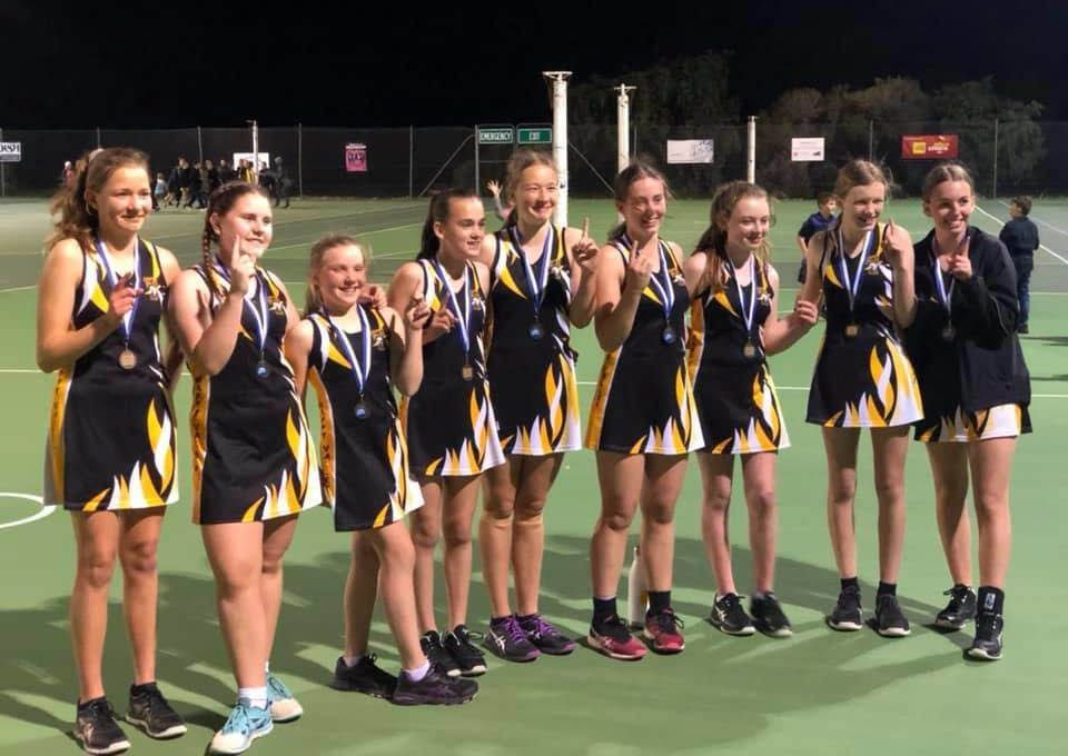 Field hockey team in dark uniforms celebrating with medals on a turf field, with a coach beside them