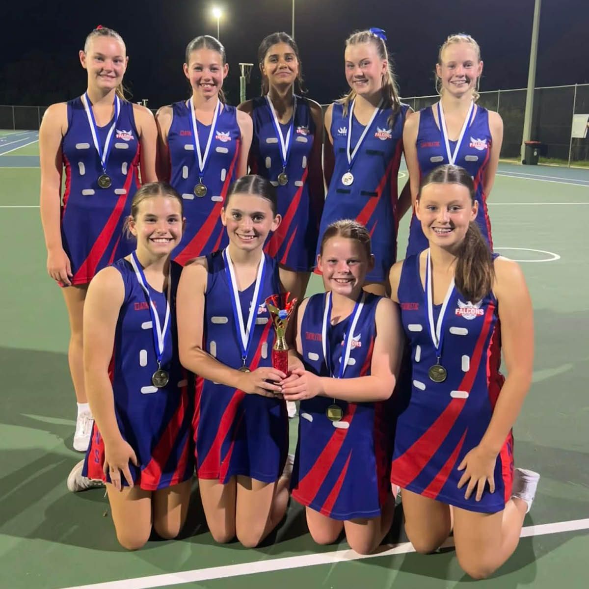 Seven medal-winning netball players in blue-red uniforms pose on a nighttime sports court.