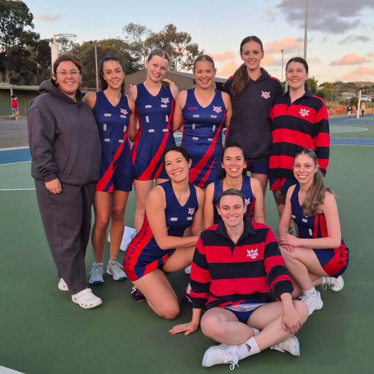 Group of netball players and a coach posing on a netball court in team uniforms at sunset.