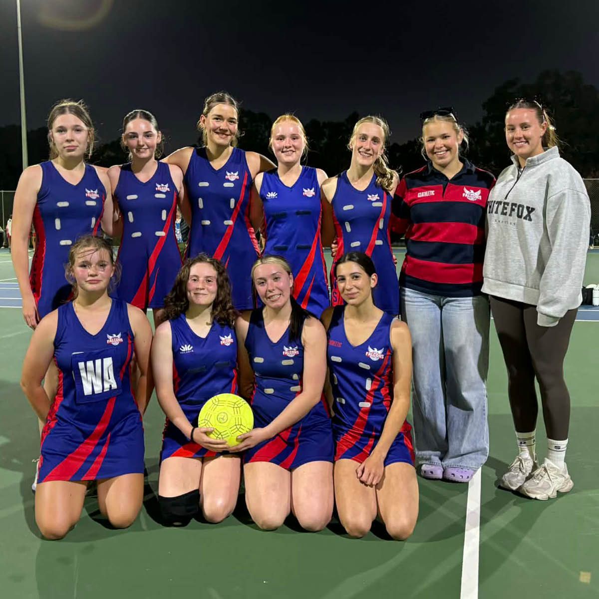 Netball team posing on an outdoor court at night, wearing blue and red uniforms, one holding a yellow ball