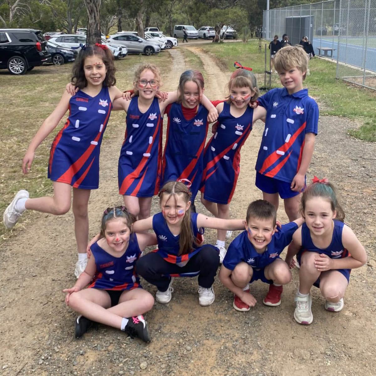 Youth sports team posing outdoors in matching blue and red uniforms, smiling on a dirt path.