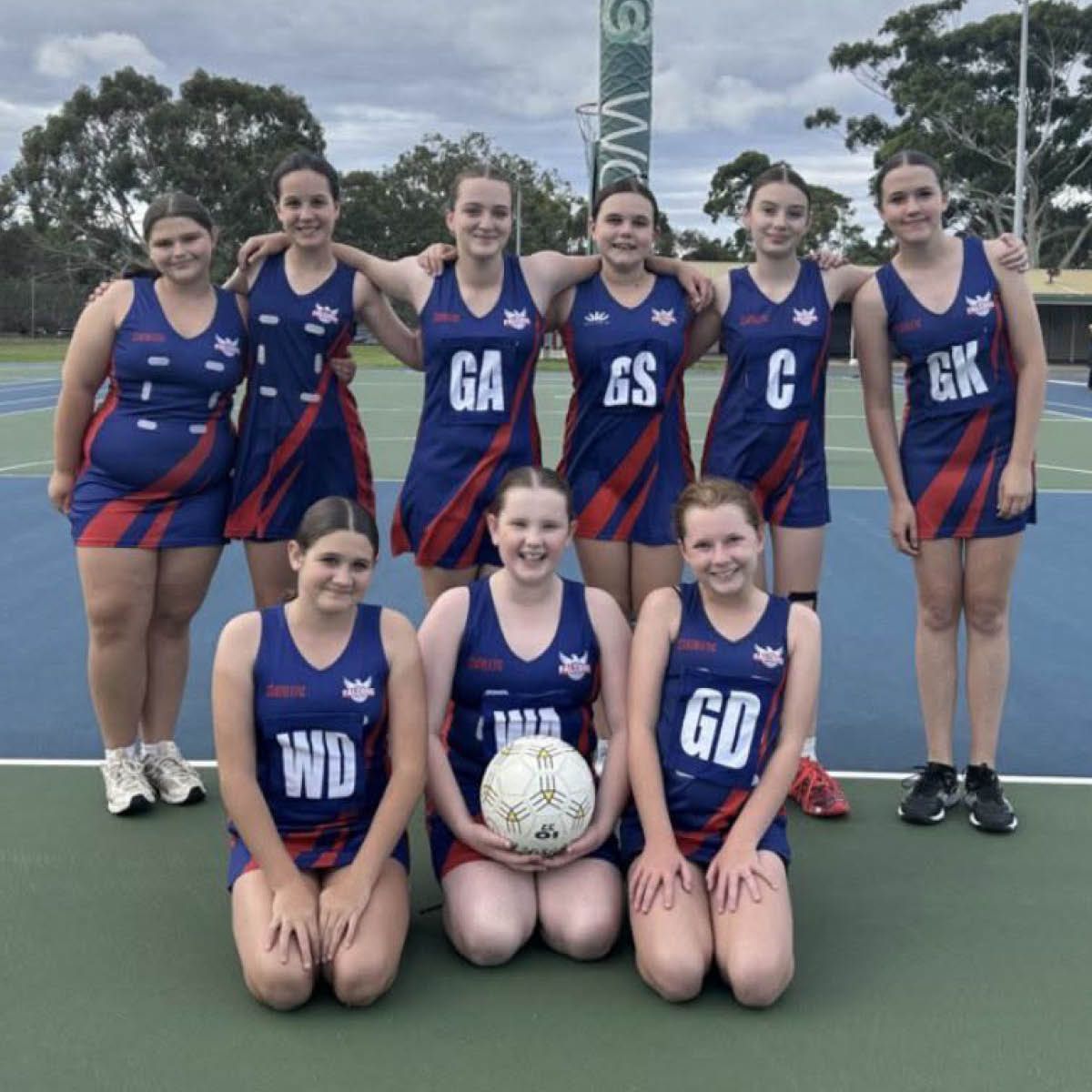 Netball team posing on an outdoor court in blue and red uniforms, with a ball centered in front.
