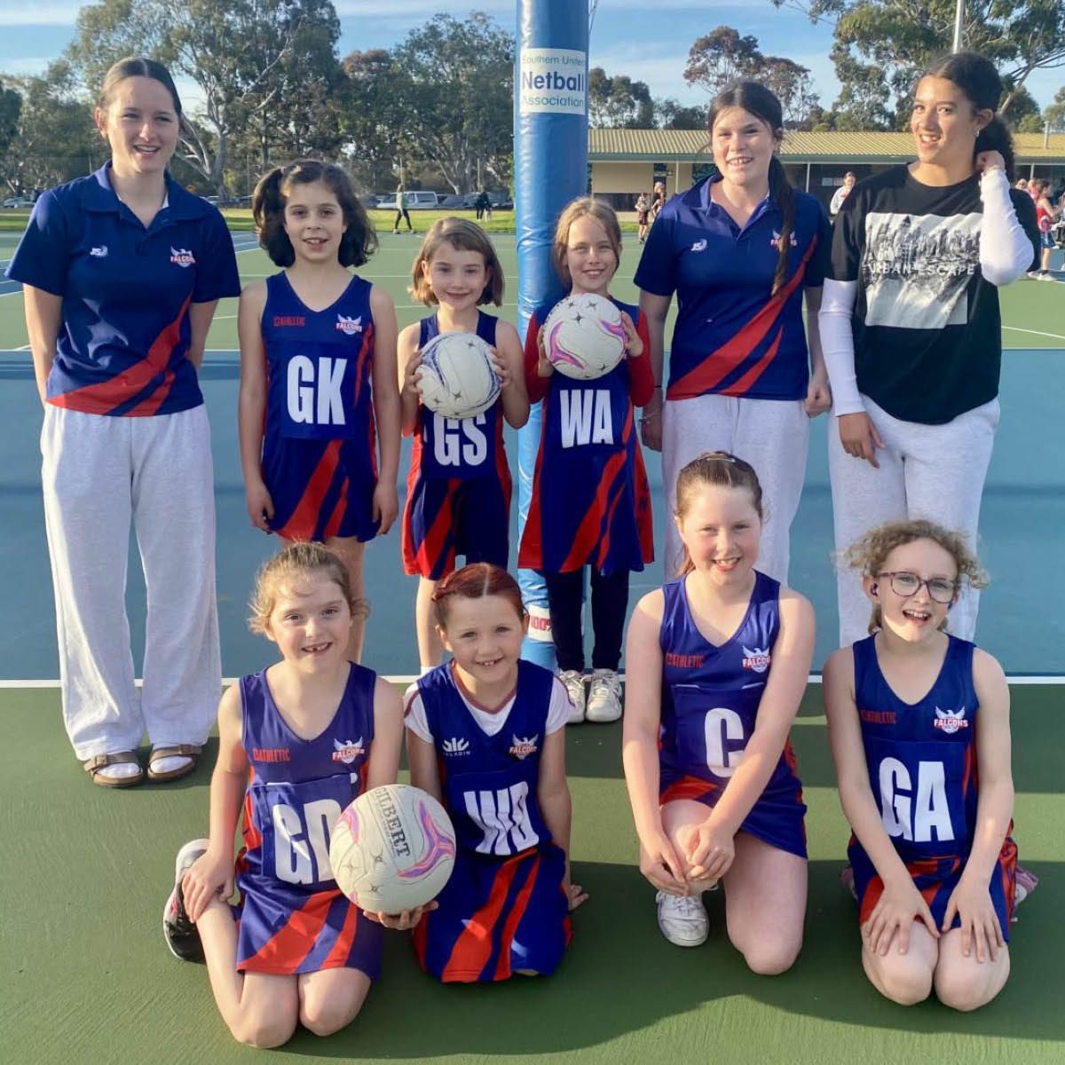 Youth netball team posing outdoors in blue-red uniforms with balls and position bibs on a sunny court