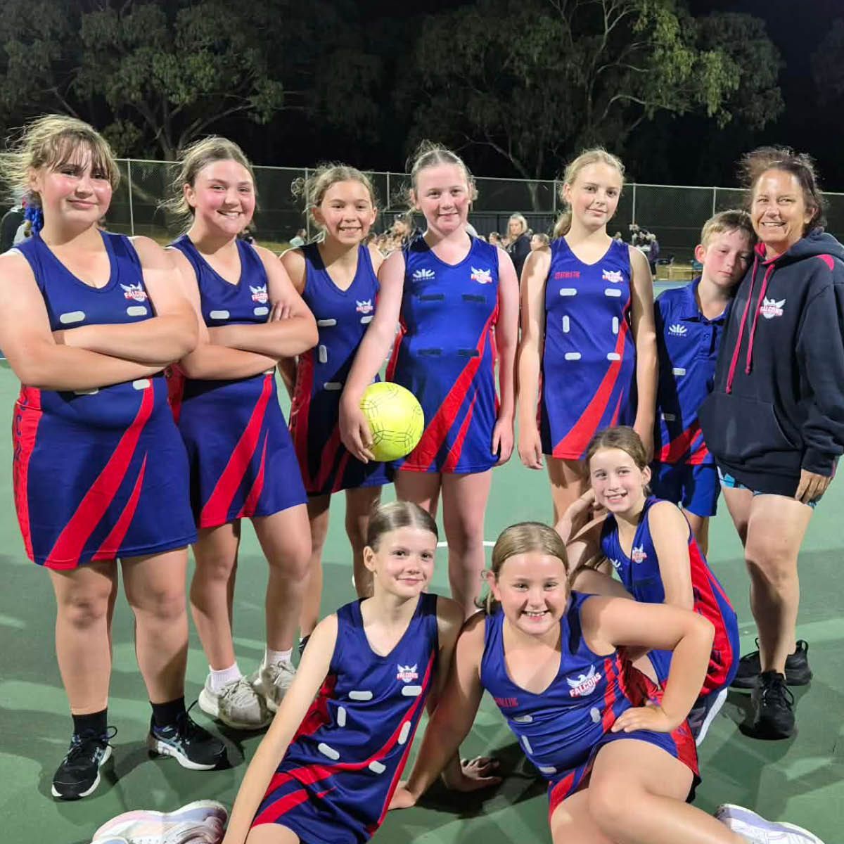 Seven netball players in blue-and-red uniforms pose on a court at night with a coach.