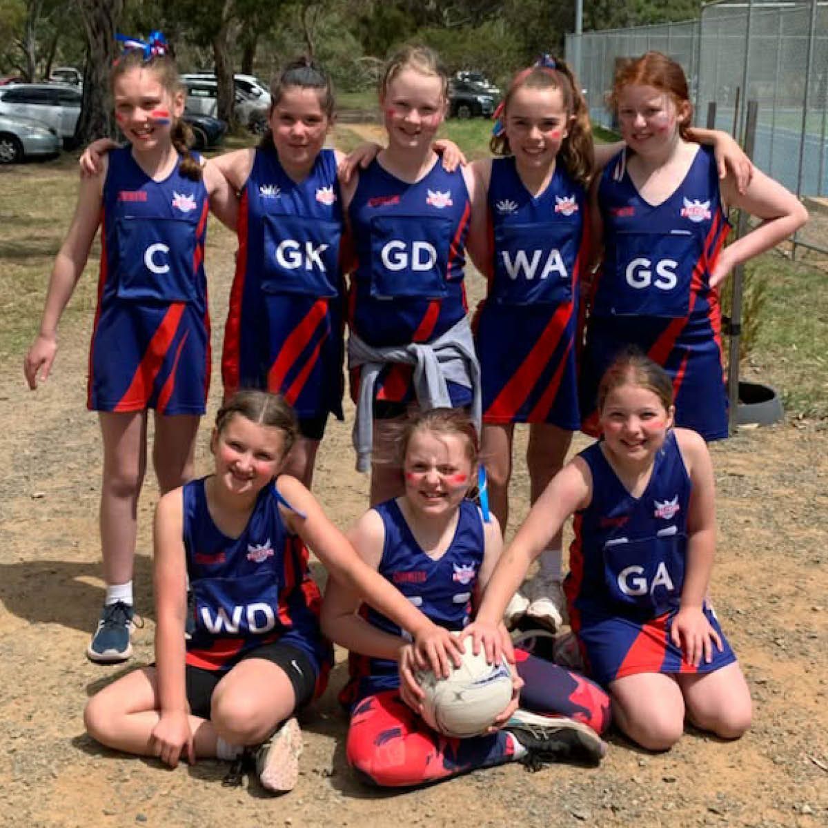 Seven netball players in blue and red uniforms pose outdoors, with two seated holding a ball.