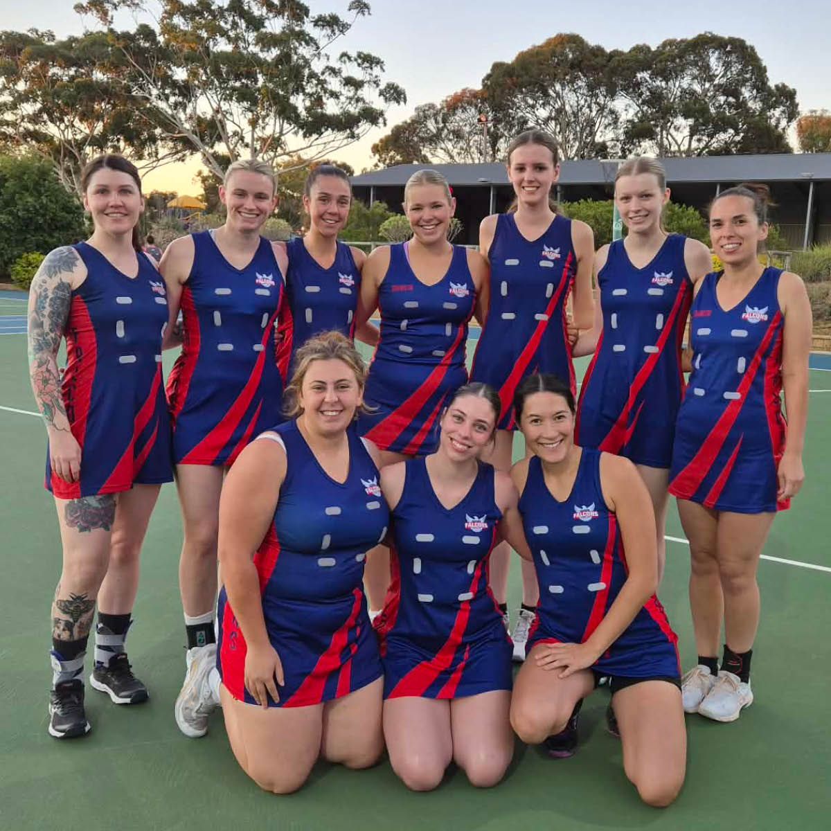 Nine women in blue and red sports uniforms posing together on an outdoor court at sunset