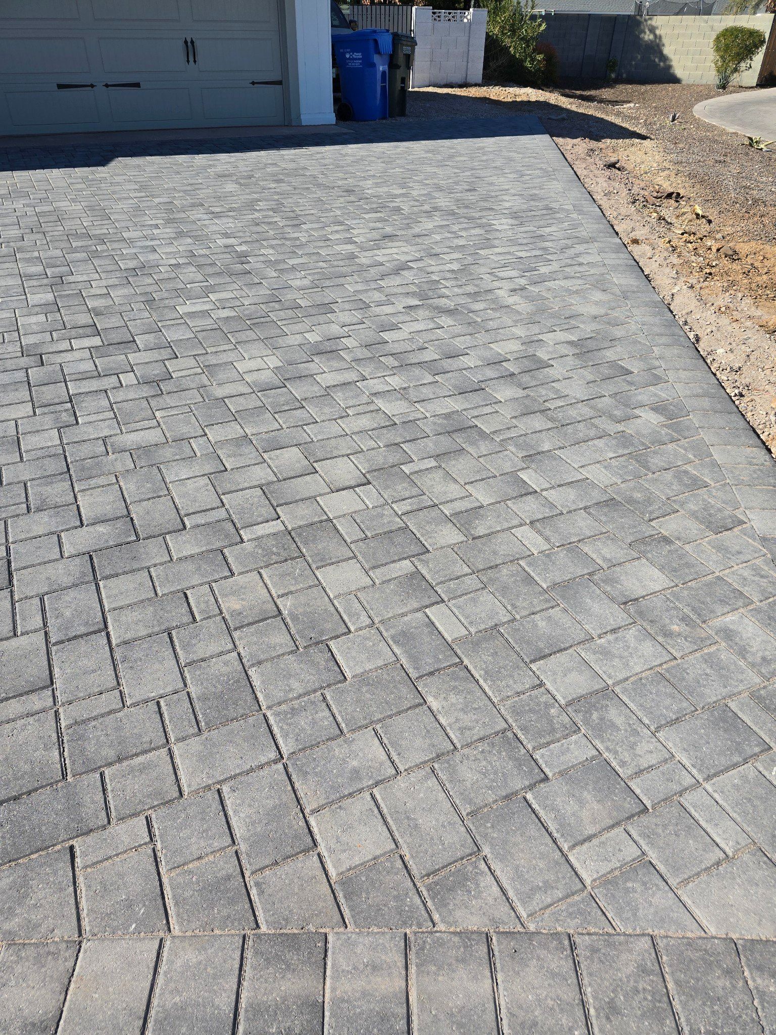 A brick driveway leading to a garage with a blue trash can in the background.