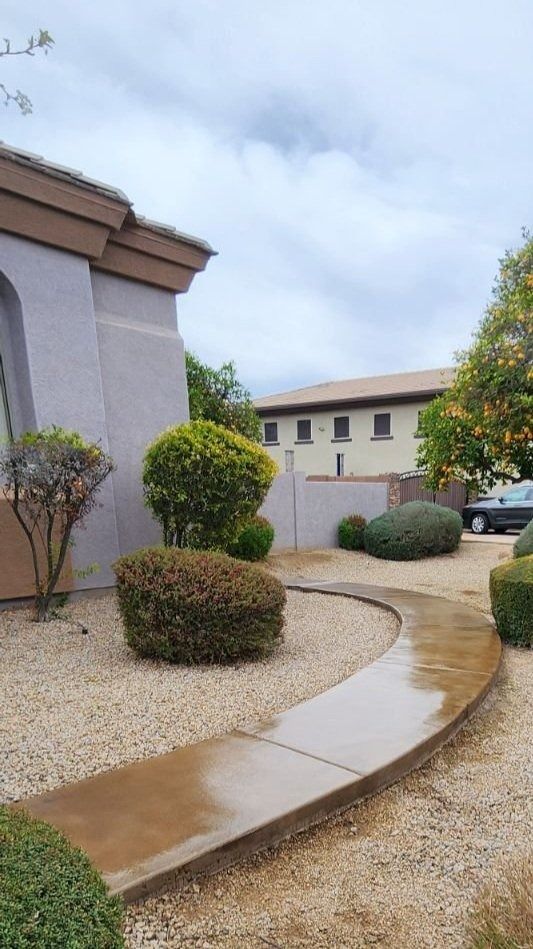 A concrete walkway leading to a house surrounded by bushes and gravel.
