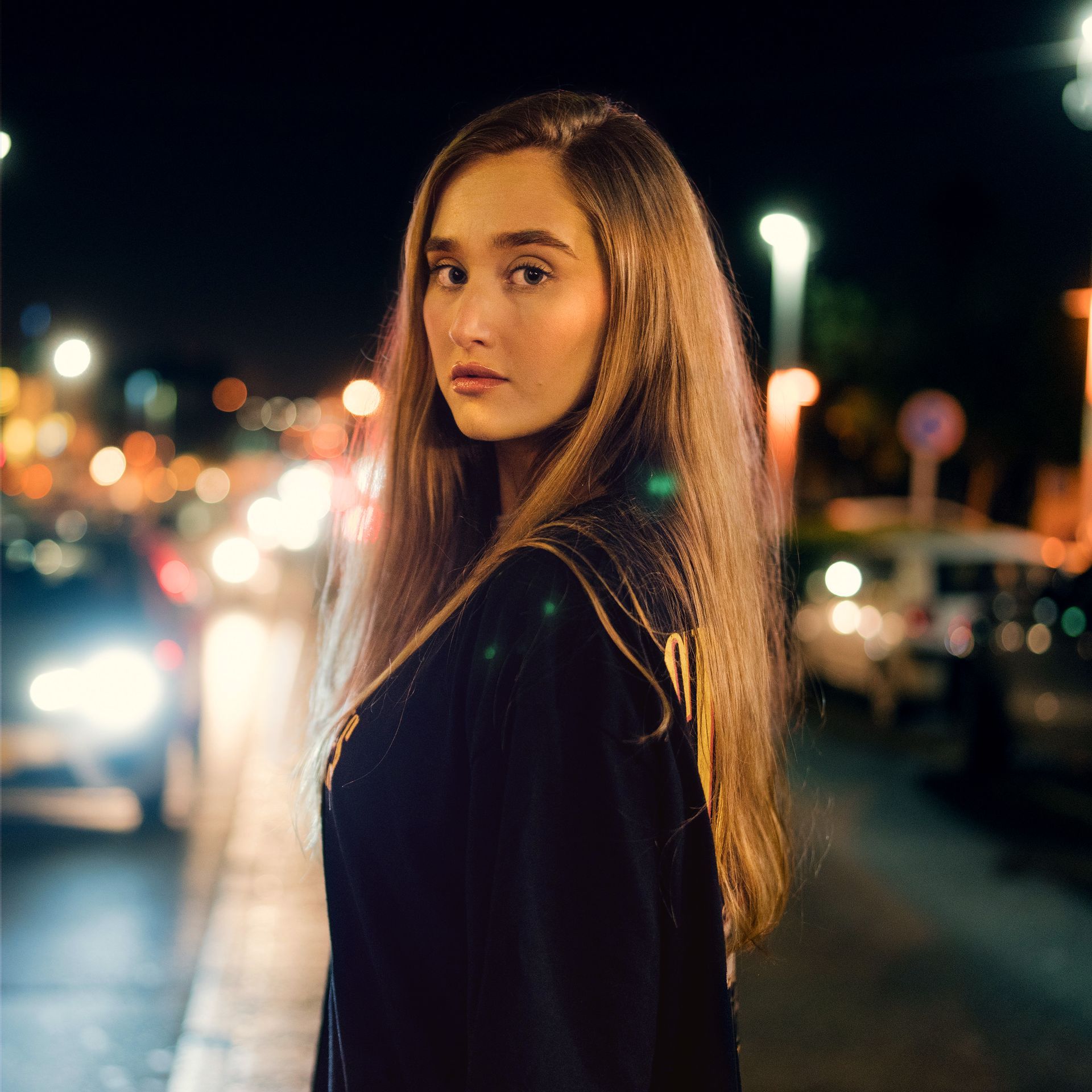 A woman with long hair is standing on a sidewalk at night