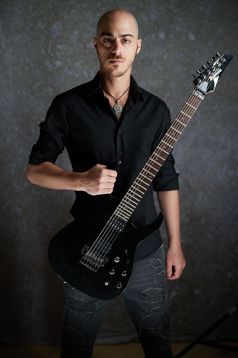 A bald man in a black shirt is holding a black electric guitar.