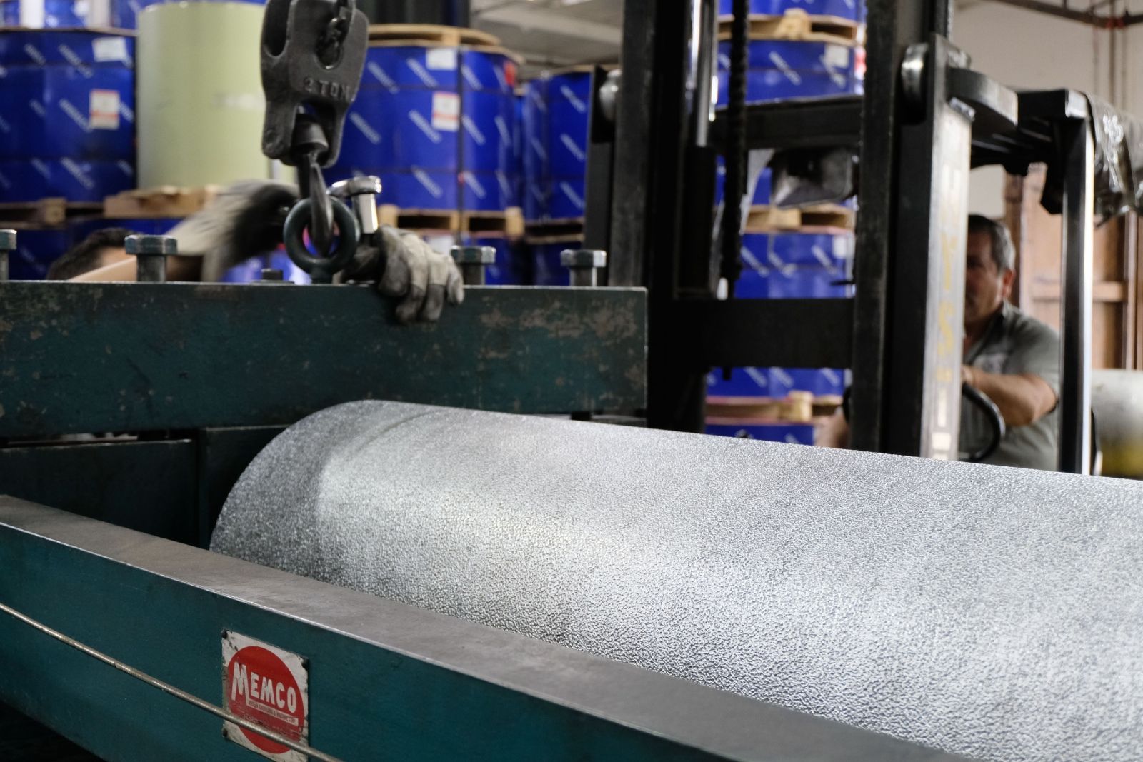 Worker operating a forklift, moving a large roll of gray material in a warehouse setting.