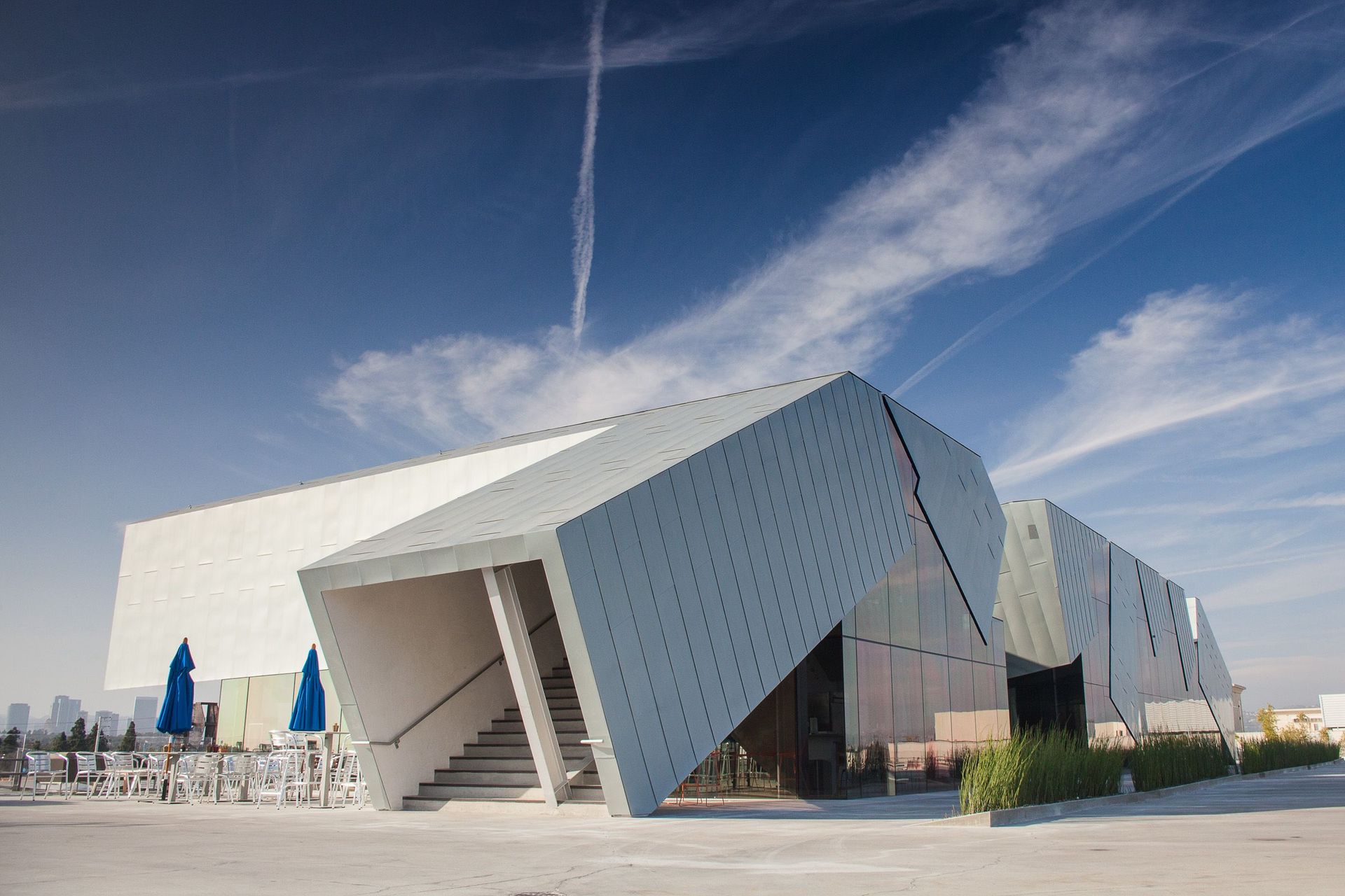 Modern white building with angled facade, stairs, blue umbrellas, and a blue sky.