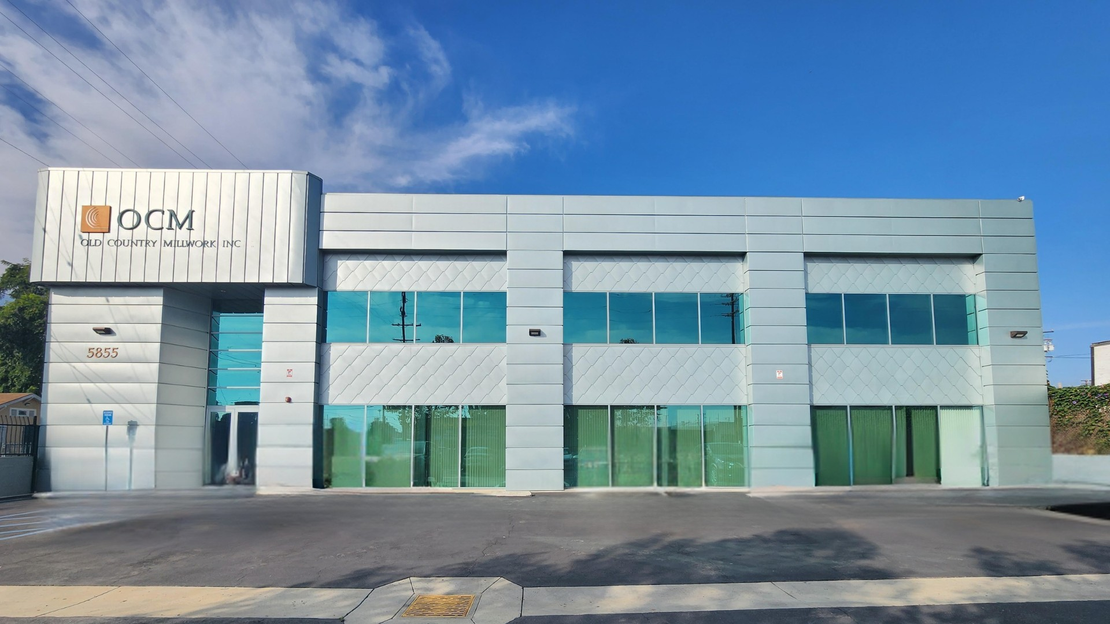 Gray building with orange logo, small bush, and metal fence on a sunny day.