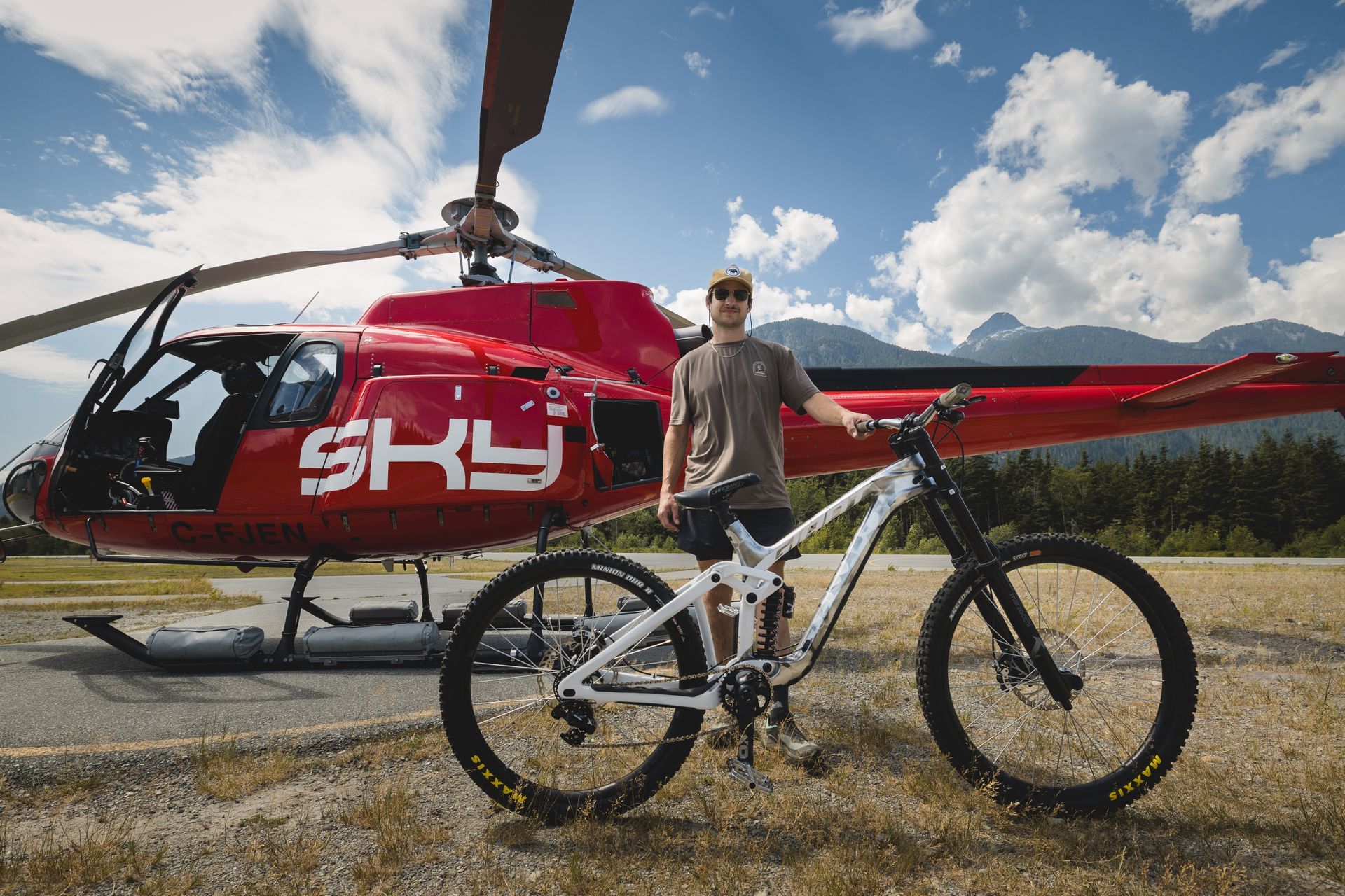 Man with mountain bike poses in front of red helicopter. Mountains and blue sky background.
