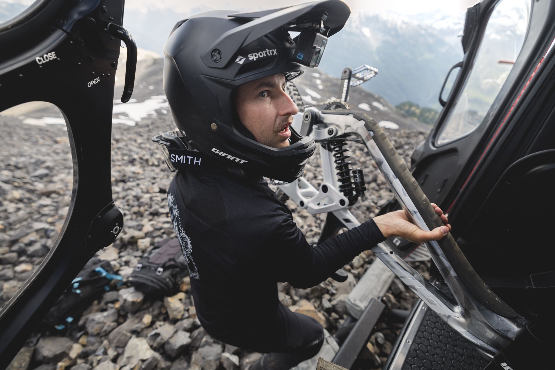 Man in helmet exits helicopter with mountain bike, rocky summit backdrop.