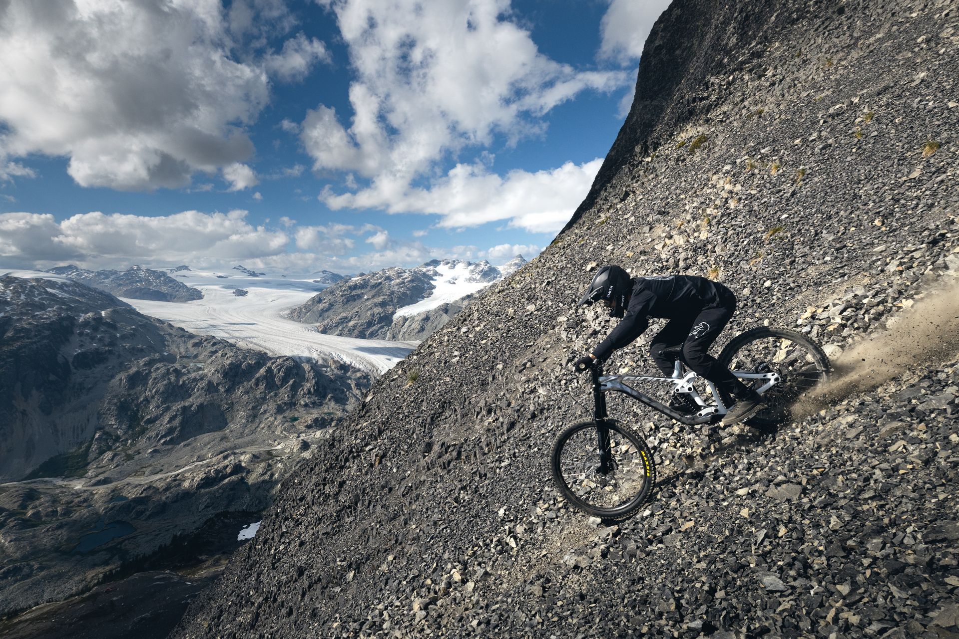 Mountain biker riding down a rocky slope, with a glacial valley in the background under a cloudy sky.