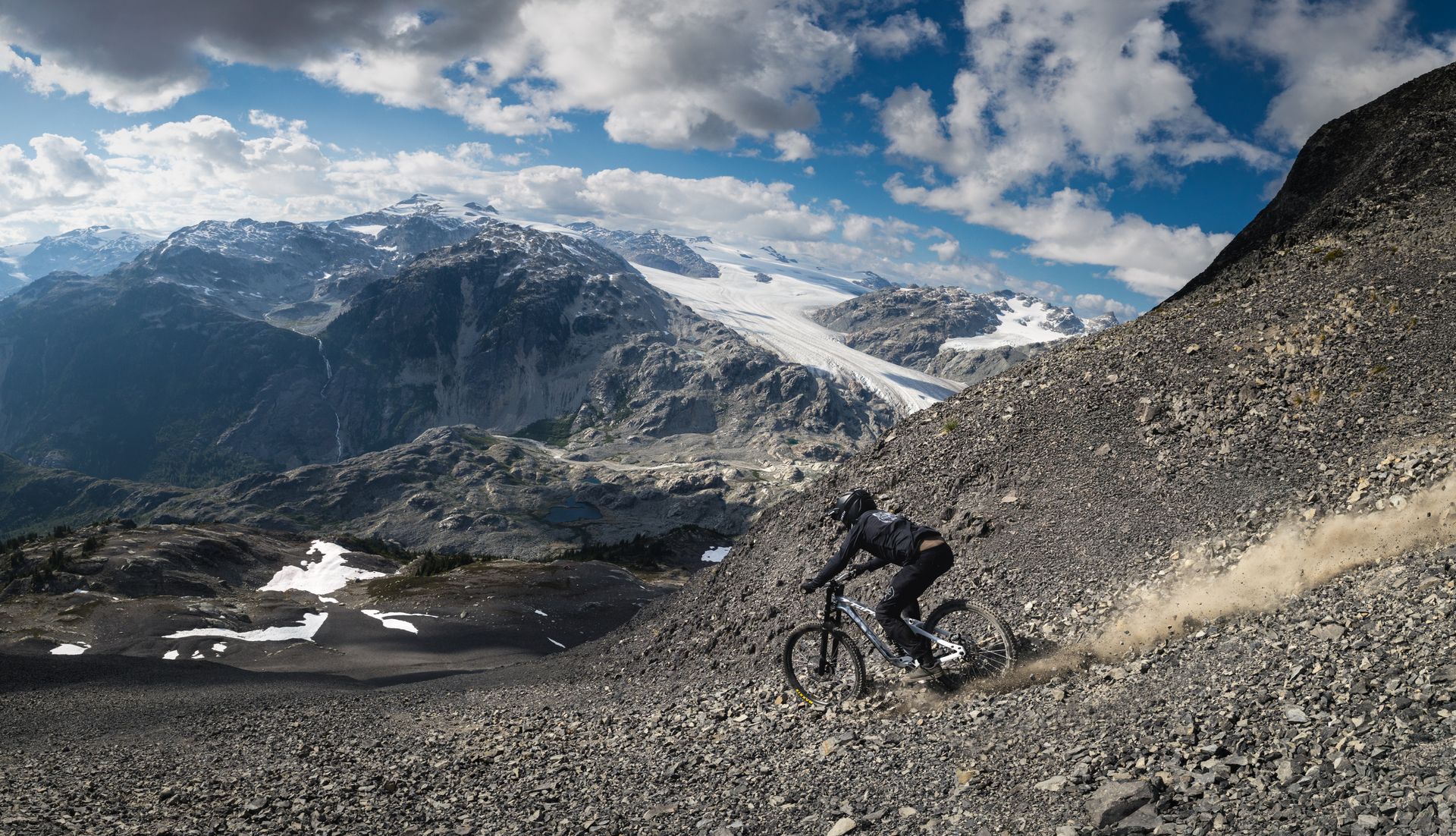 Mountain biker descends a rocky slope with snow-capped peaks and blue sky in the background.