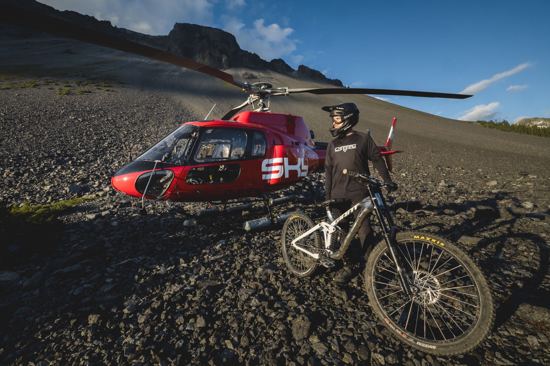 A person with a mountain bike stands beside a red helicopter on a rocky mountain slope.