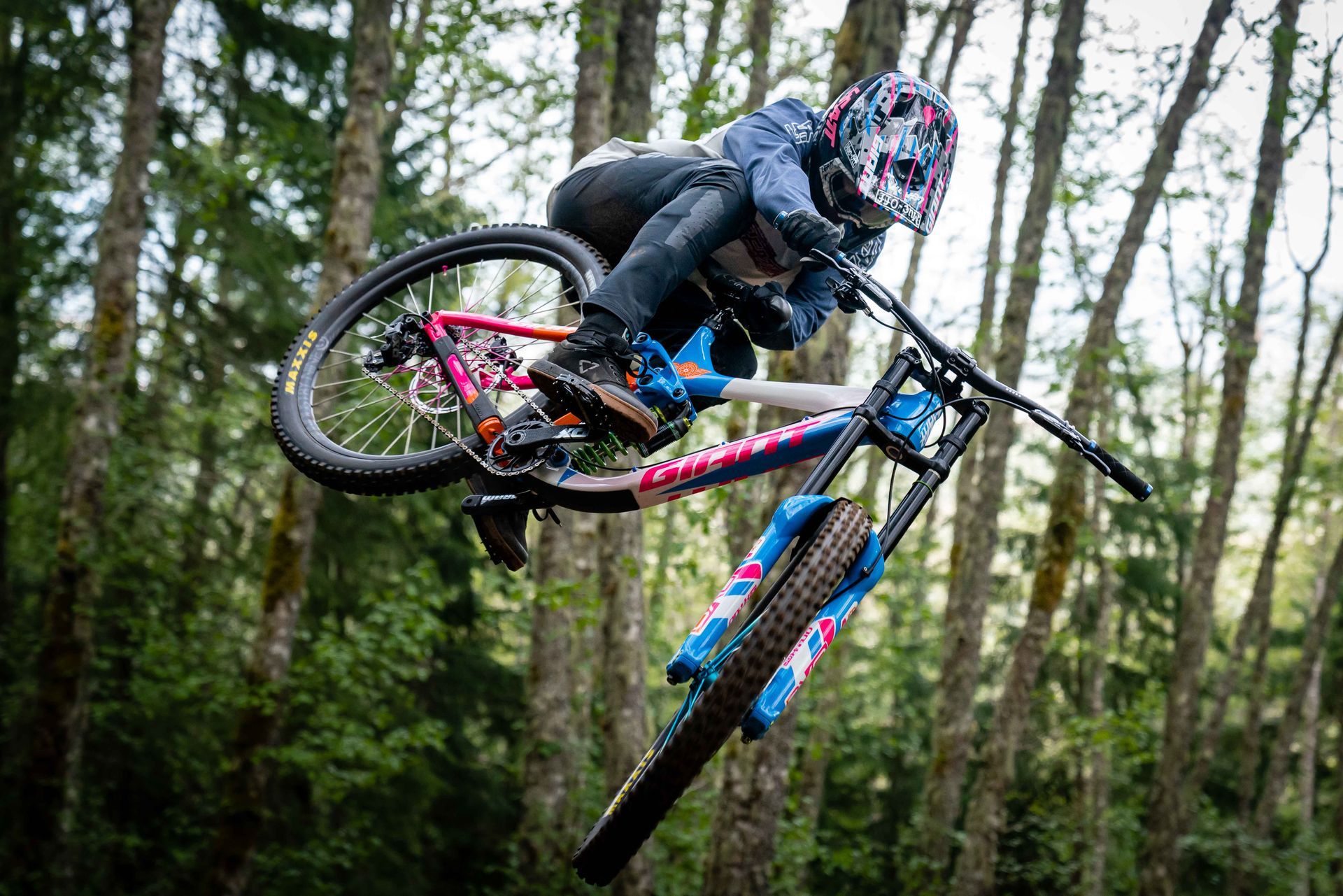 Mountain biker in mid-air, performing a trick on a trail, surrounded by trees.