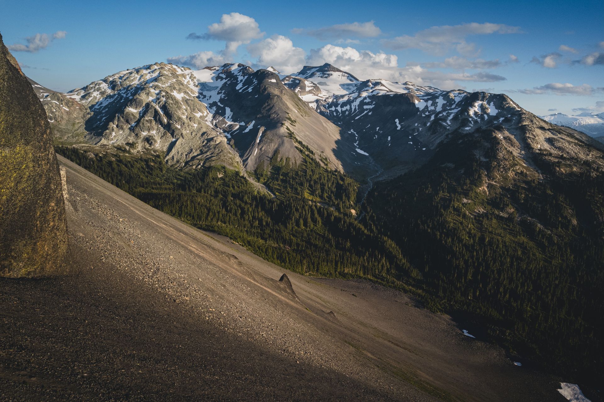 Mountain range with rocky slopes, evergreen trees, and patches of snow under a blue sky.