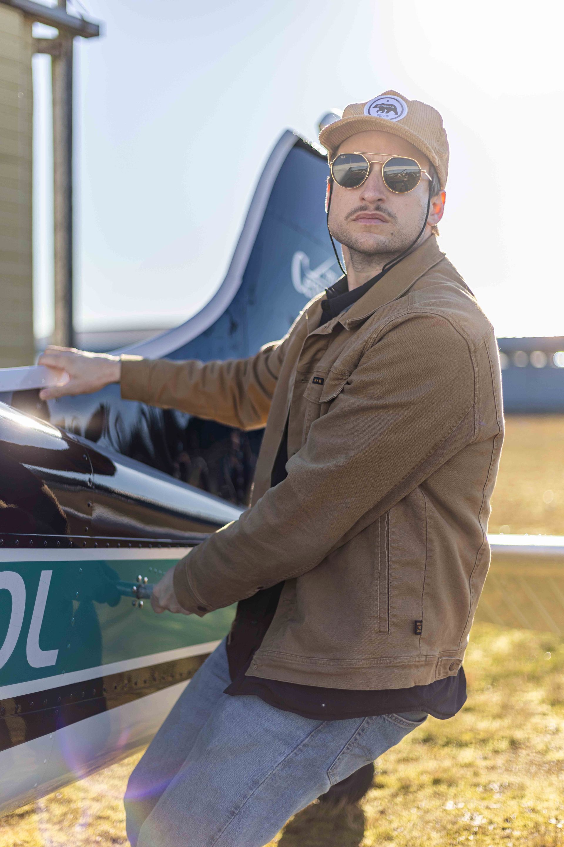 Man in sunglasses and jacket near a small airplane, holding the wing. Bright sunlight.