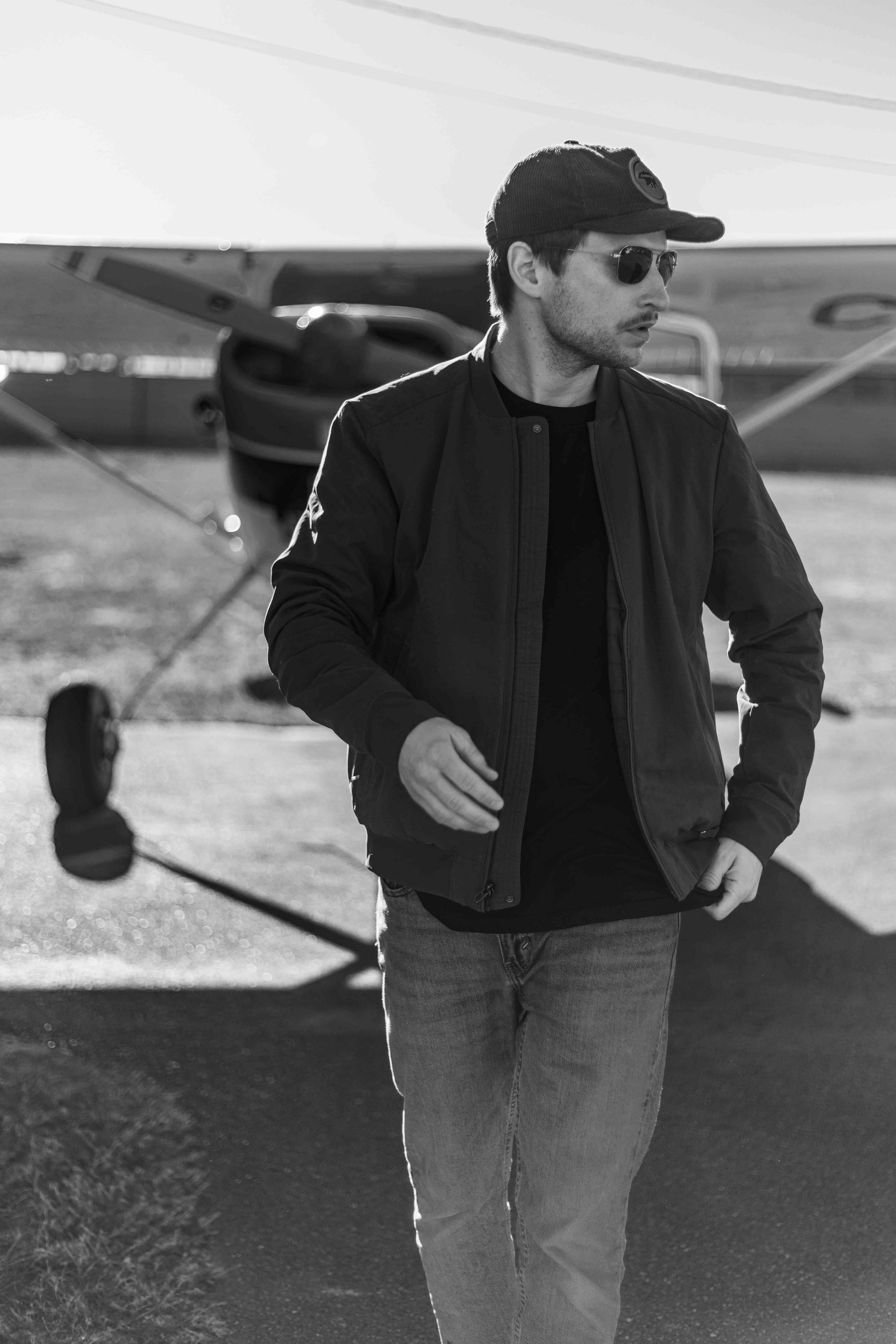 Man in bomber jacket and cap stands near a small airplane on a sunny airfield.