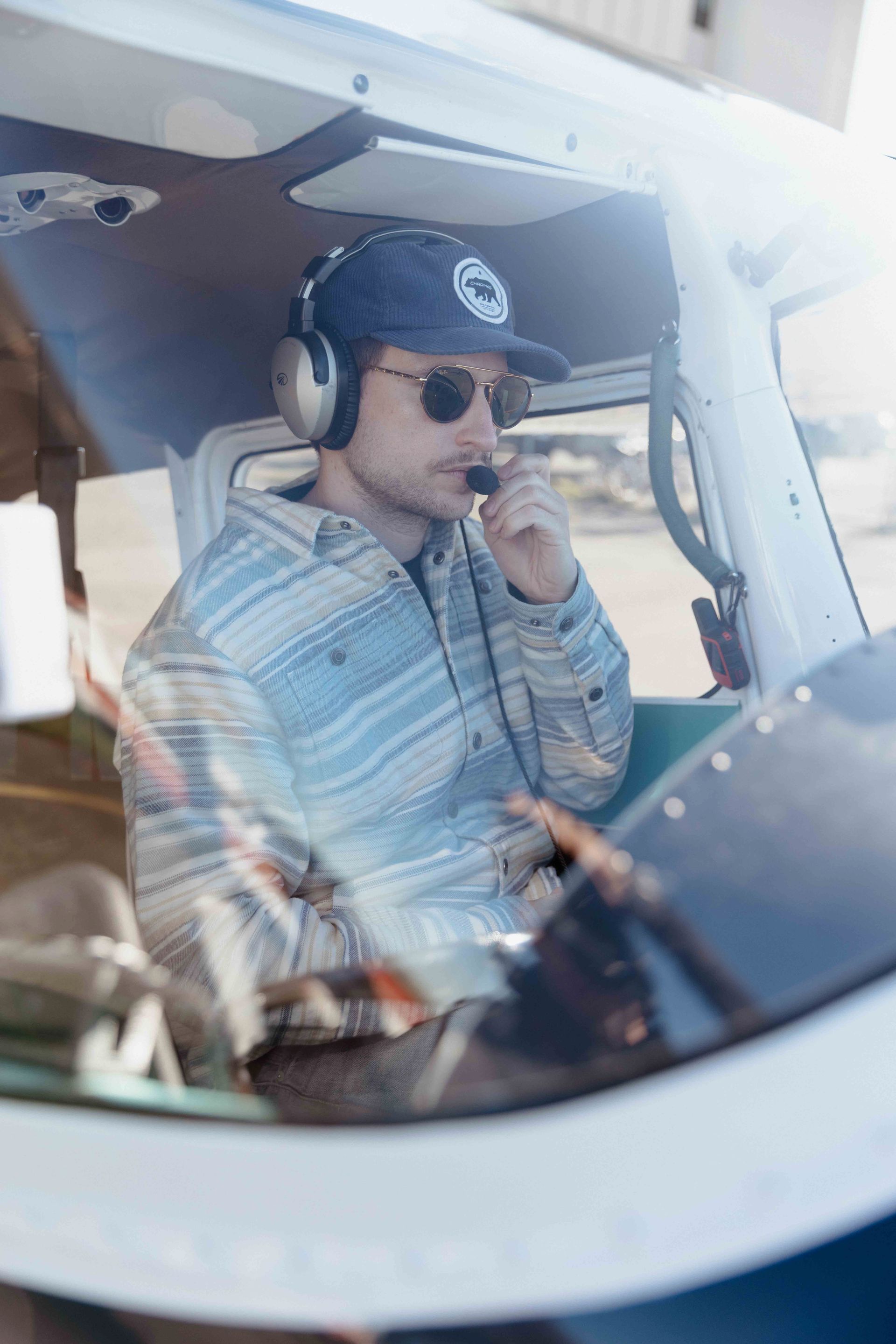 Man in a small airplane cockpit wearing sunglasses, hat, and headset, talking on a microphone.