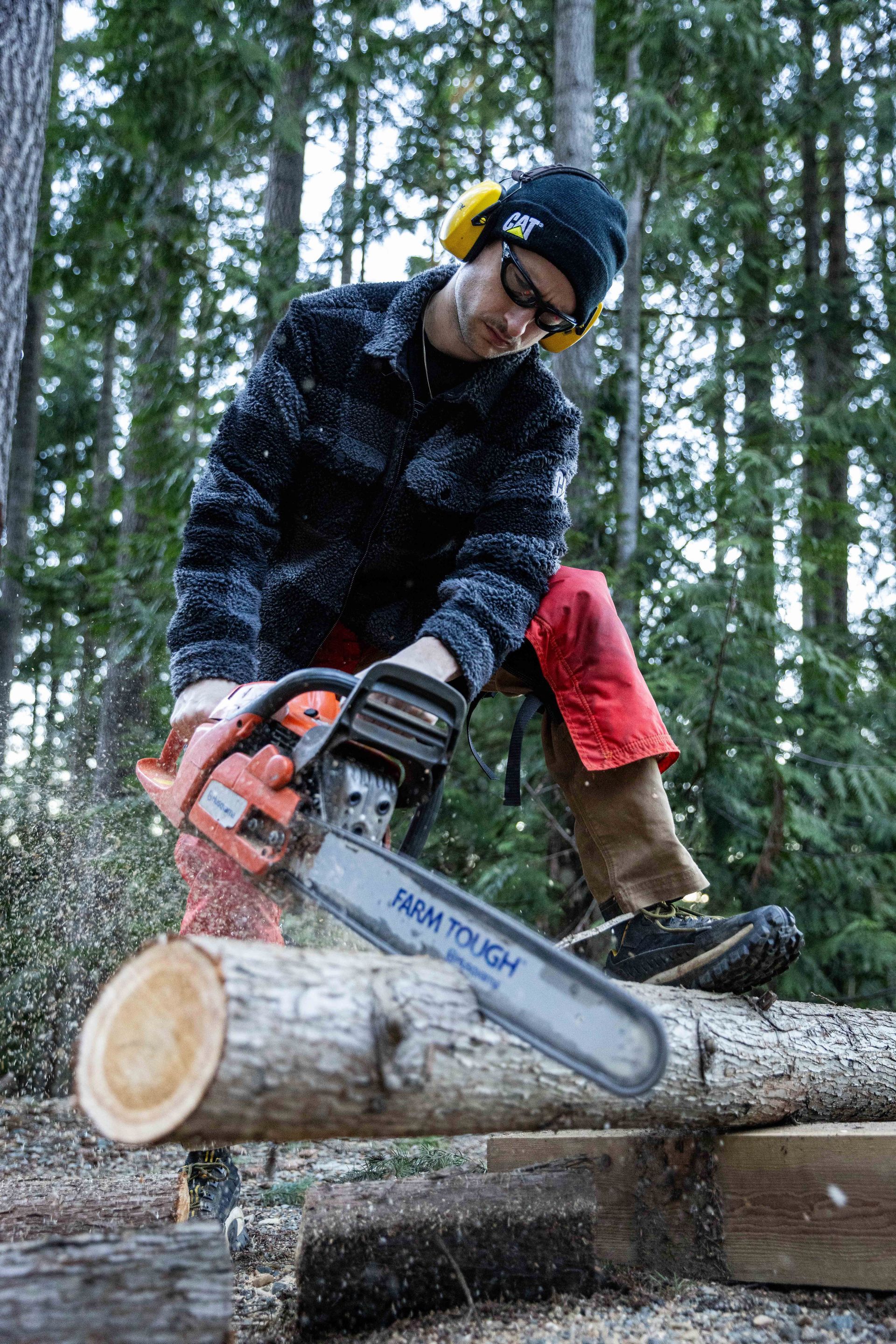 Person wearing safety gear cutting a log with a chainsaw in a forest.