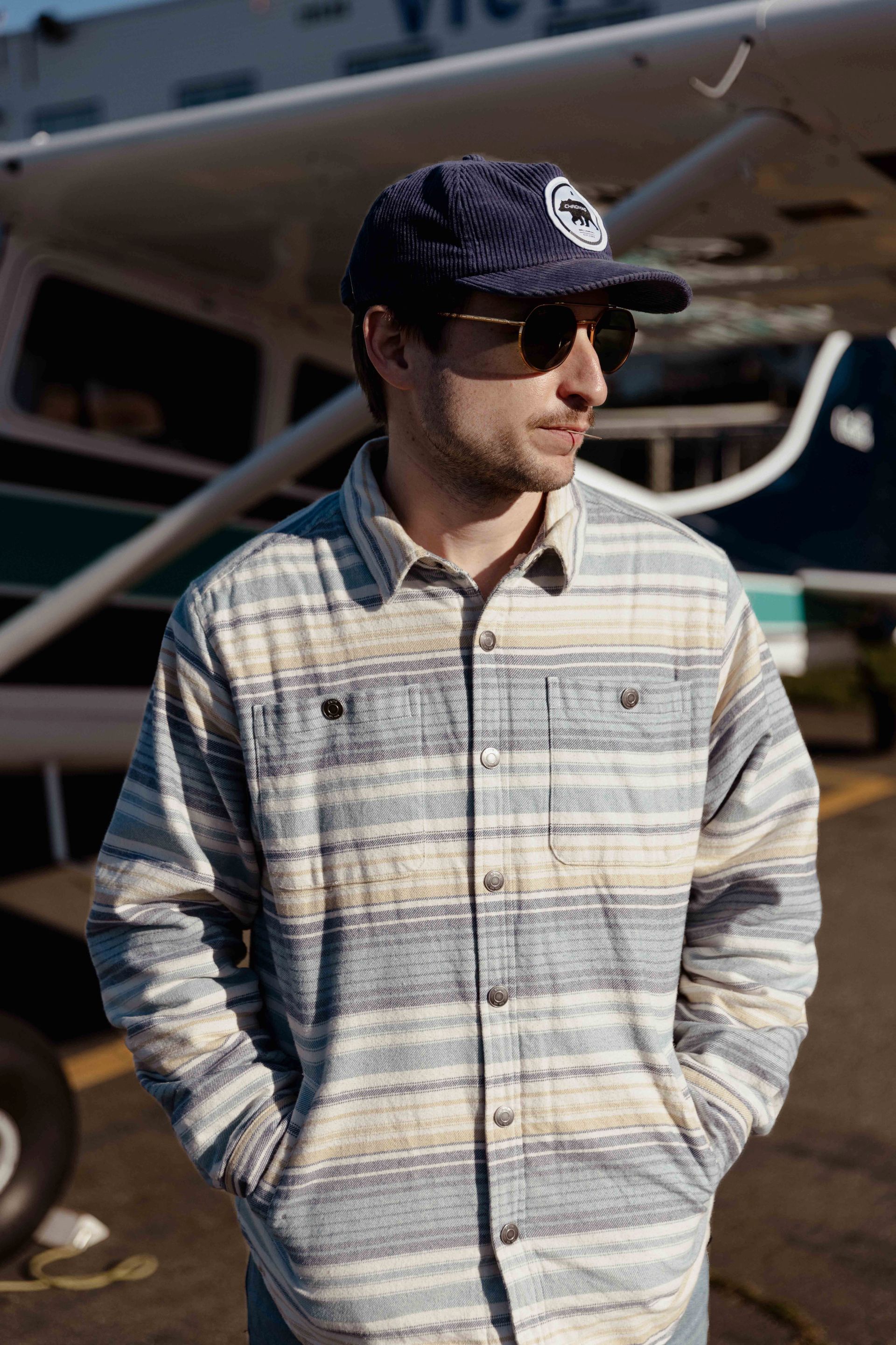 Man wearing sunglasses, hat, and striped jacket stands near a small airplane.